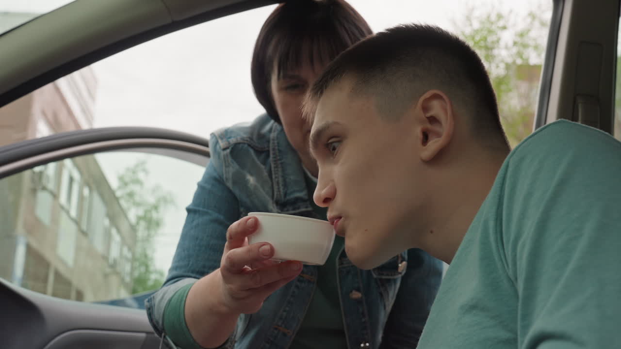 White Teen Being Fed From Caregiver Cup Inside Parked Car, Mutual Smiles And Trust, Bowl And Cup Handover, Nurturing Support, Roadside Compassionate Care And Shared Moment Of Nourishment