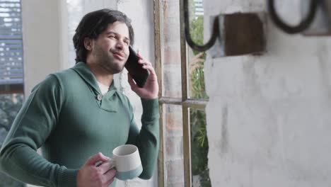 Hombre-Caucásico-Tomando-Café,-Hablando-Por-Teléfono-Inteligente-Y-Mirando-Por-La-Ventana-En-Casa,-Cámara-Lenta