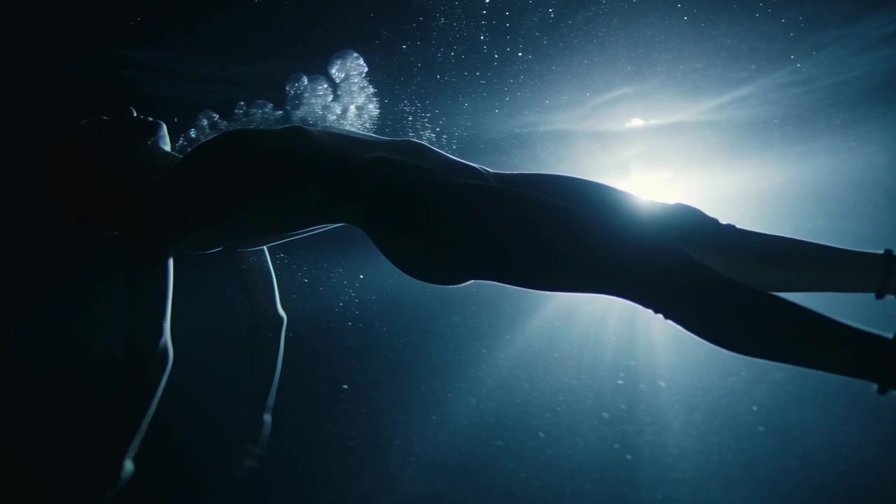 Mulher flutuando de maiô levantando os braços, exalando bolhas em direção à luz de fundo na piscina, com partículas iluminadas.