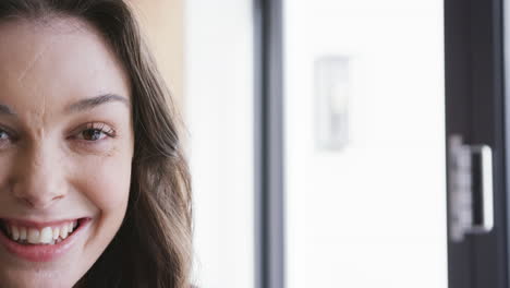 Half-face-of-happy-caucasian-woman-standing-by-window-and-smiling-in-sunny-home,-copy-space