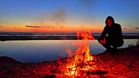 Slow-motion-landscape-view-of-person-kneeling-by-campground-site-with-fire-hot-burning-embers-next-to-lake-river-bay-sunset-camping-adventure-travel