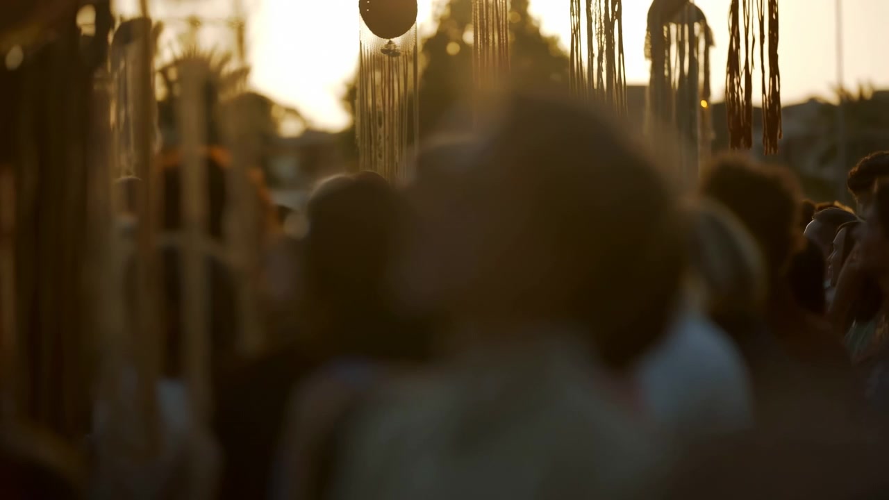Teste e spalle sfocate riempiono la parte inferiore della scena, mentre lunghe nappe fibrose e ornamenti rotondi pendono dall'alto in un caldo bagliore del tramonto, creando un forte contrasto, texture e un'atmosfera festosa.