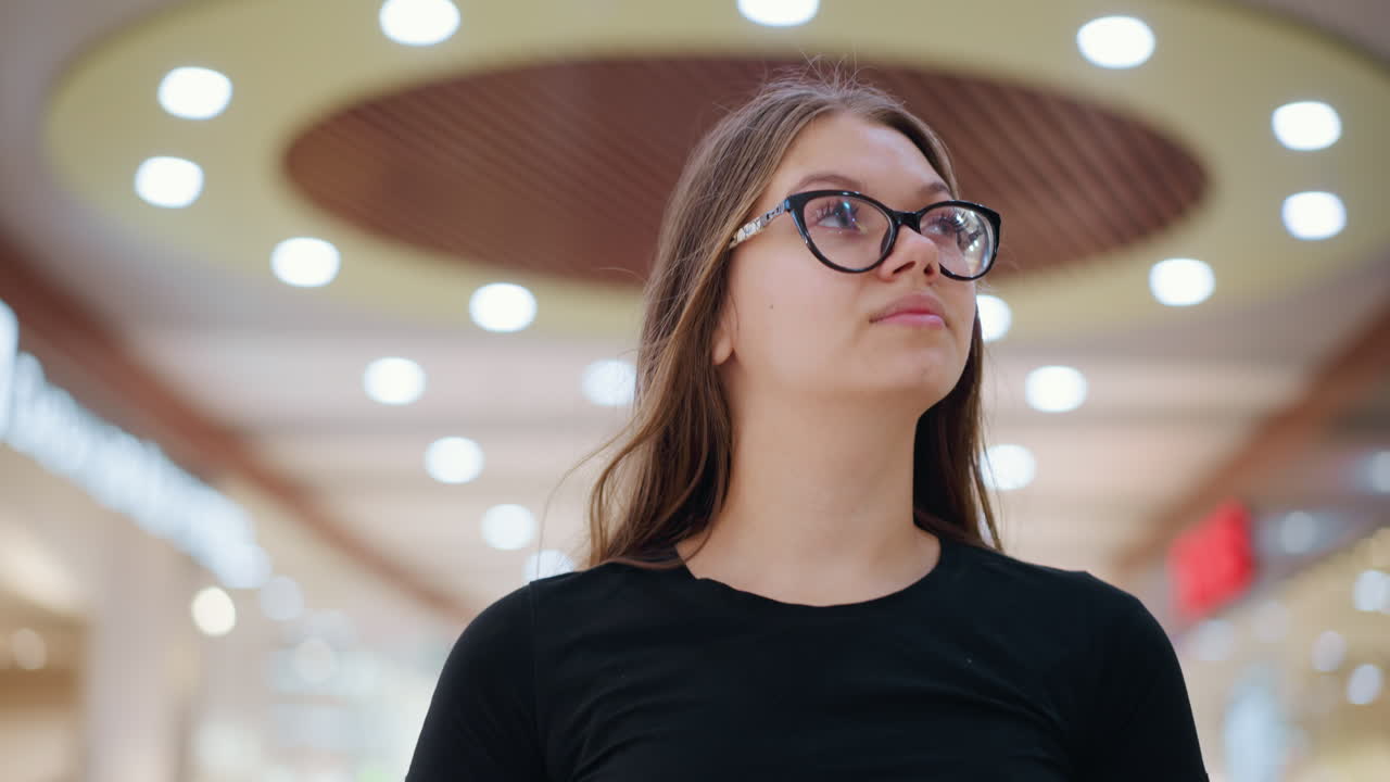 joven con camiseta negra y gafas se centra en algo en el fondo con luces borrosas y estructuras indistintas, creando una expresión reflexiva en un entorno interior moderno