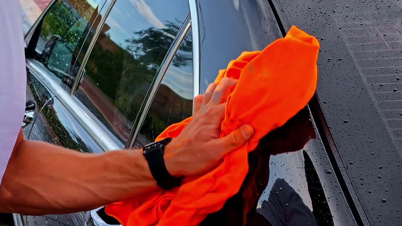 In a slow-motion close-up, a man's hand carefully dries a clean, wet black car with a bright orange microfiber towel after washing, representing auto detailing and car care