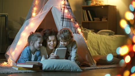 loving mother and two little spending time together in cozy room at home and reading fairy tale with tablet in teepee tent at night