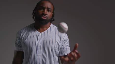 Studio-Portrait-Of-Serious-Male-Baseball-Player-Wearing-Team-Shirt-Holding-Bat-And-Throwing-Ball-In-The-Air-Shot-Against-Grey-Background-1