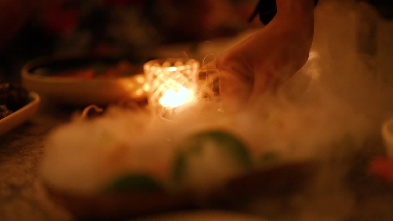 A hand interacts with a smoky dish in a dimly lit setting, creating a warm and atmospheric dining experience