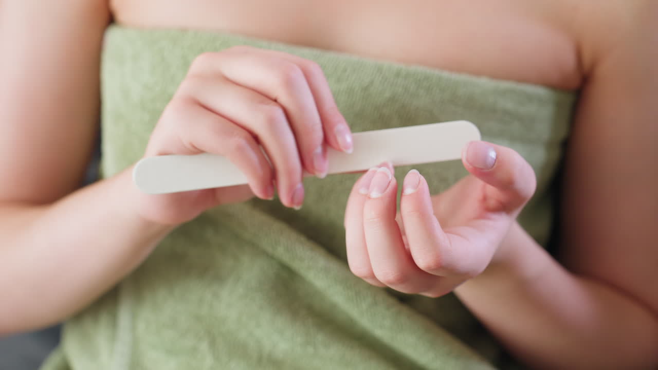 Close up hand view of woman gently filing fingernails using white nail filer while wrapped in green towel, nails well-shaped, focus on selfcare and grooming in natural indoor light