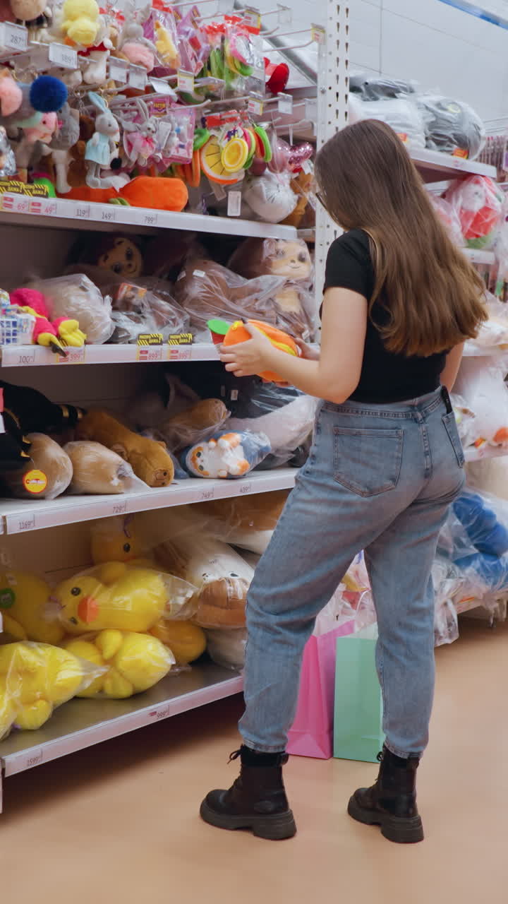 joven con camiseta negra sostiene e inspecciona un vibrante bolso de peluche naranja con detalles verdes en un centro comercial bien iluminado, examina cuidadosamente el artículo mientras navega por los estantes llenos de juguetes coloridos