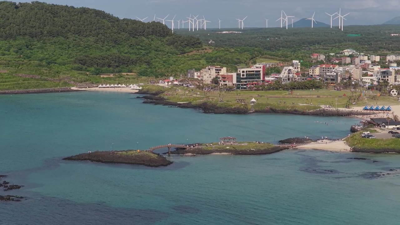 Horisontalt utsnitt som beveger seg bakover til høyre nær Hamdeok-stranden i Sinheung-ri, Jeju-øya, Sør-Korea, som viser turkist vann, steinete holmer, kystlinje og landsby