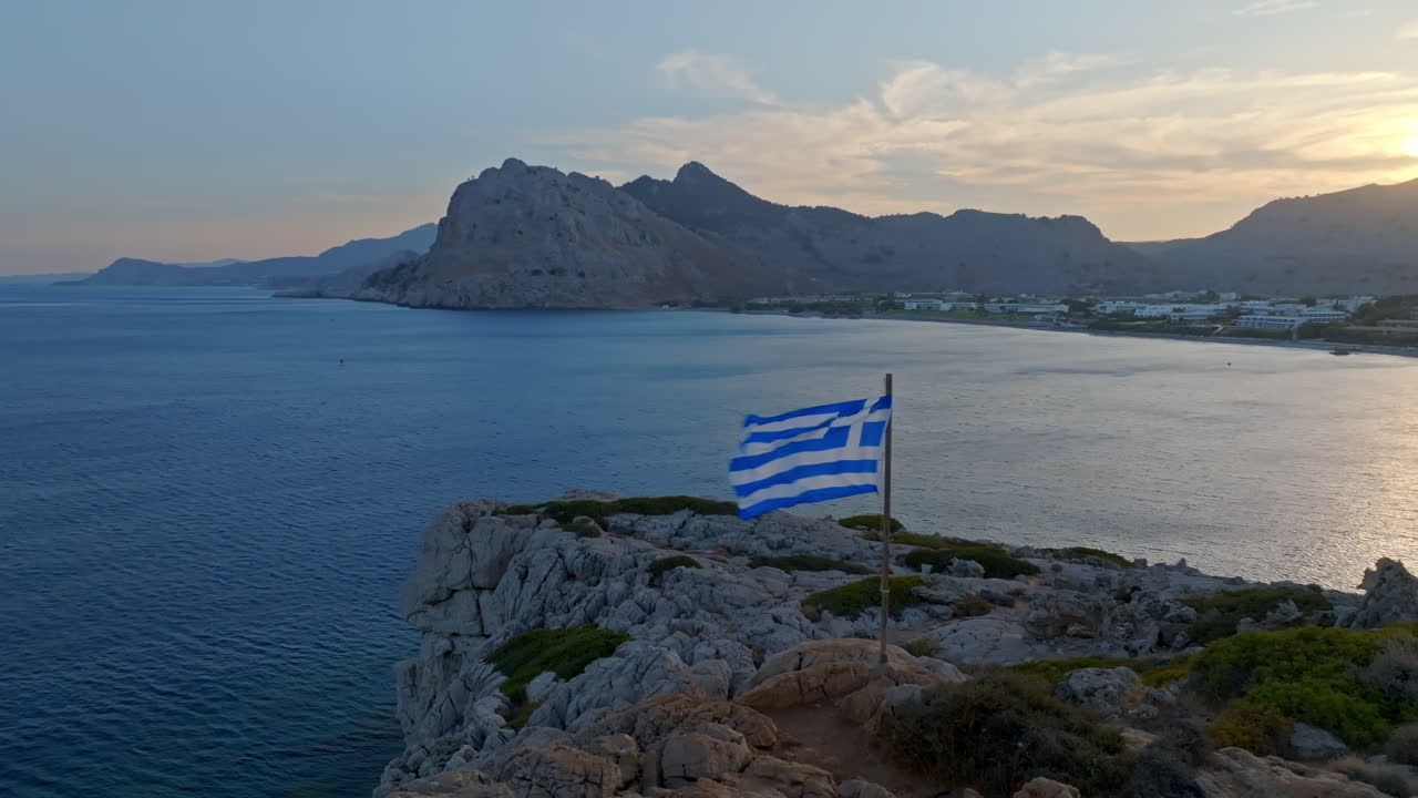 Aerial view orbiting a Greek flag on the coastline of Kolymbia, sunset in Rhodes, Greece