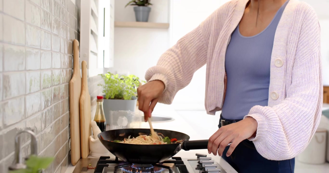 Mujer de mediana edad revolviendo fideos salteados en la cocina, generando iconos de categoría que orbitan un wok que se desvanece.