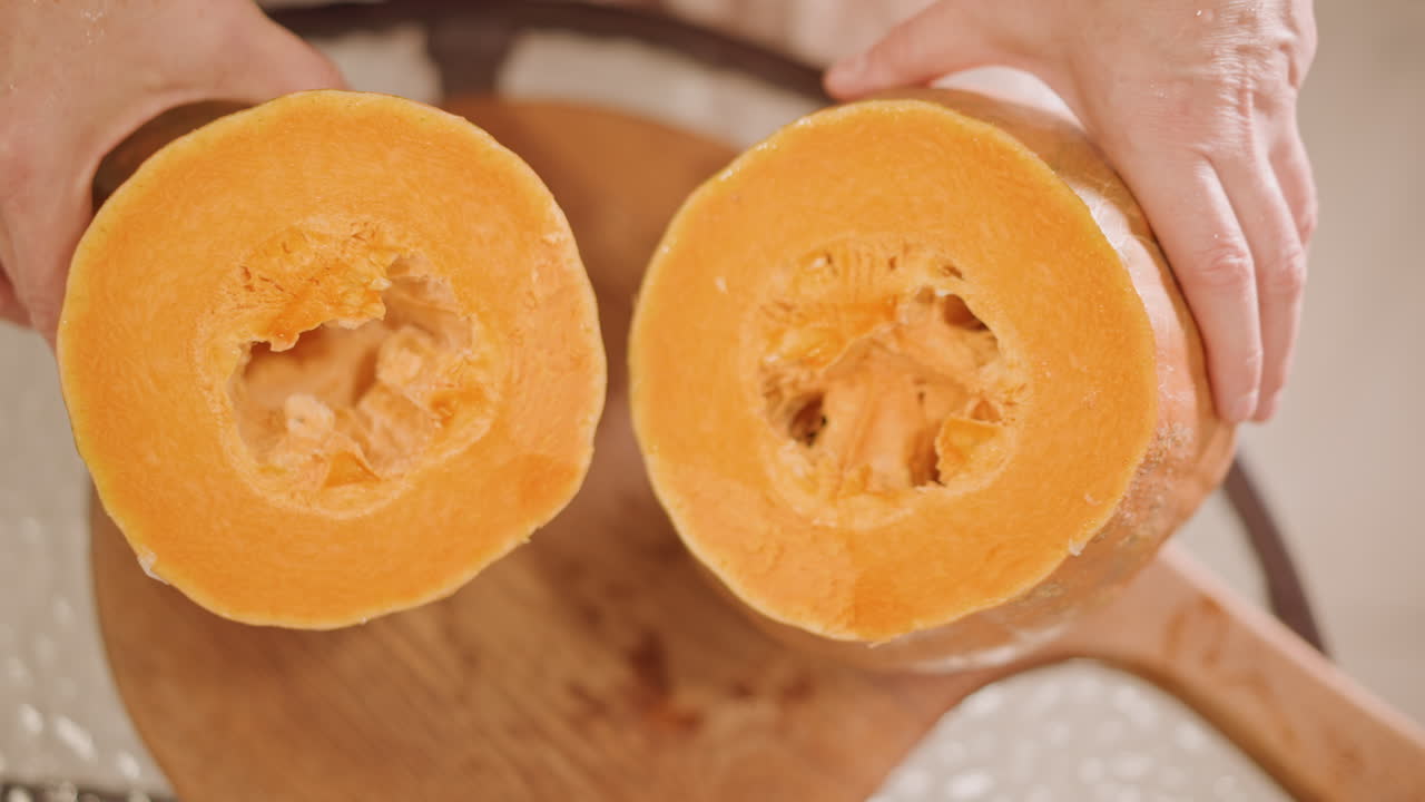 Dos calabazas partidas por la mitad sostenidas sobre una tabla, primer plano de manos que muestran mitades ahuecadas a juego, corazón fibroso visible y bolsas de semillas, preparación para relleno de tarta o asado, luz natural brillante, artesanal.