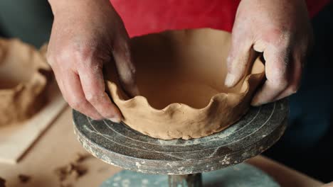 a close-up of an artisan potter's hands holding a newly crafted, rustic clay bowl with a wavy rim, presenting the finished product on a banding wheel in a workshop