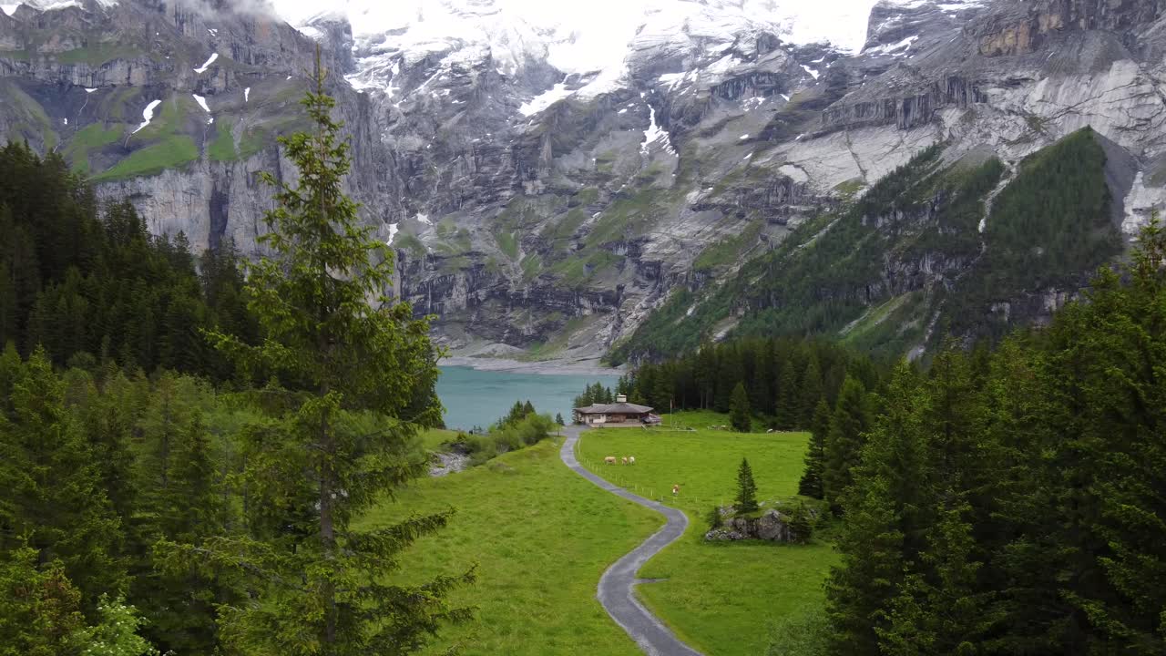 Vista aerea in ascesa di un isolato chalet in legno su un verde pascolo alpino, circondato da montagne e pini, con vista sul lago glaciale Oeschinensee, turchese e azzurro, a Kandersteg, Svizzera.