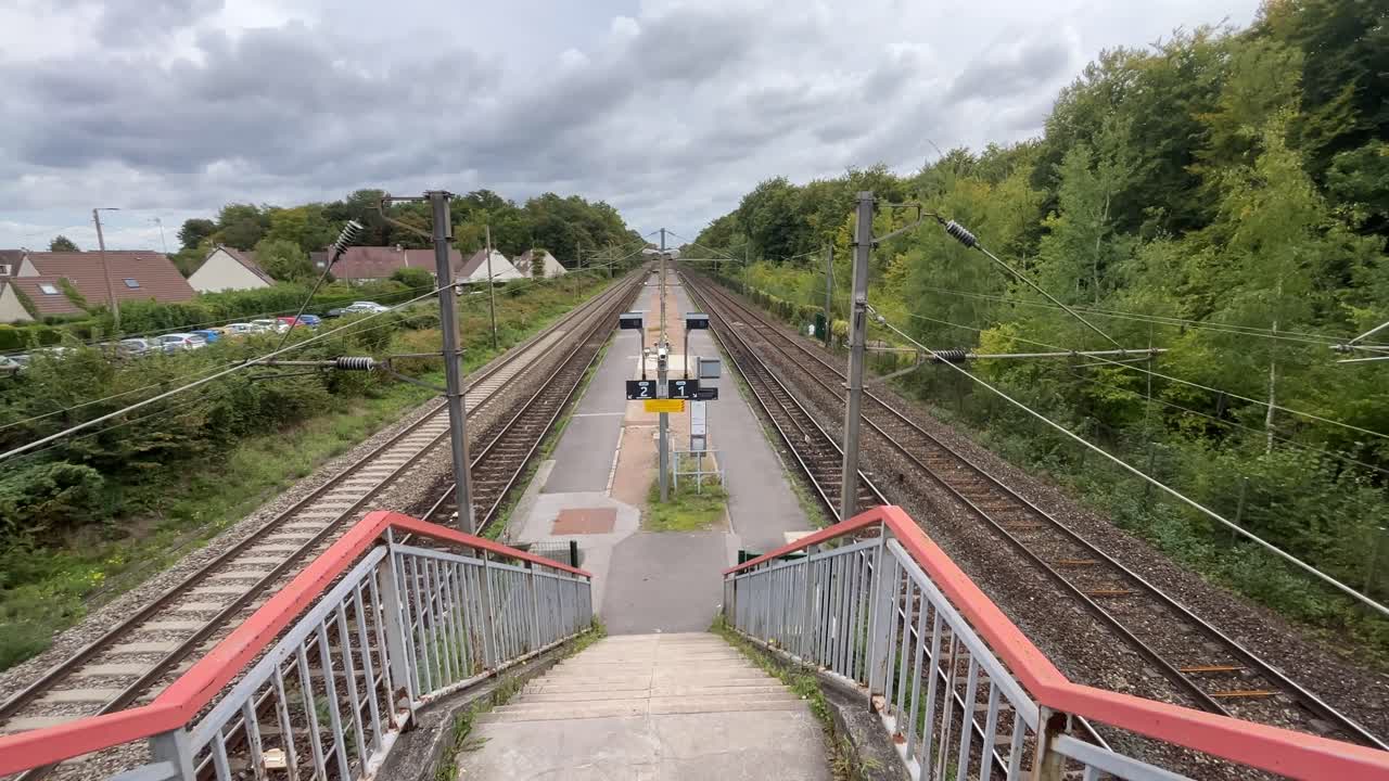 A fixed shot shows a staircase descending onto railway tracks at a small train station near Beaumont in the Paris region, with houses and vegetation around
