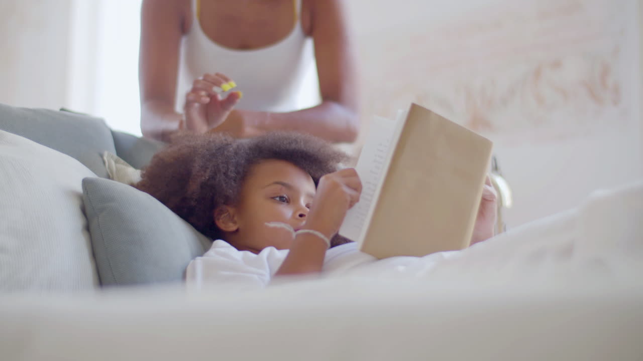 Little African American girl spending time reading in bed when her mum coming up, smiling and kissing her