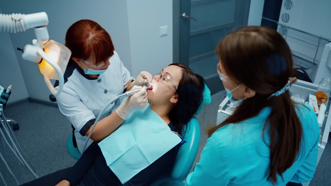 Professional female stomatologist at work. Young woman sitting in dental chair during teeth treatment. Dentist uses modern medical equipment in dentistry. Top view.