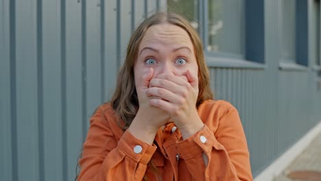 brown hair woman in orange jacket raises hands to cover mouth in shock awe fear, urban background