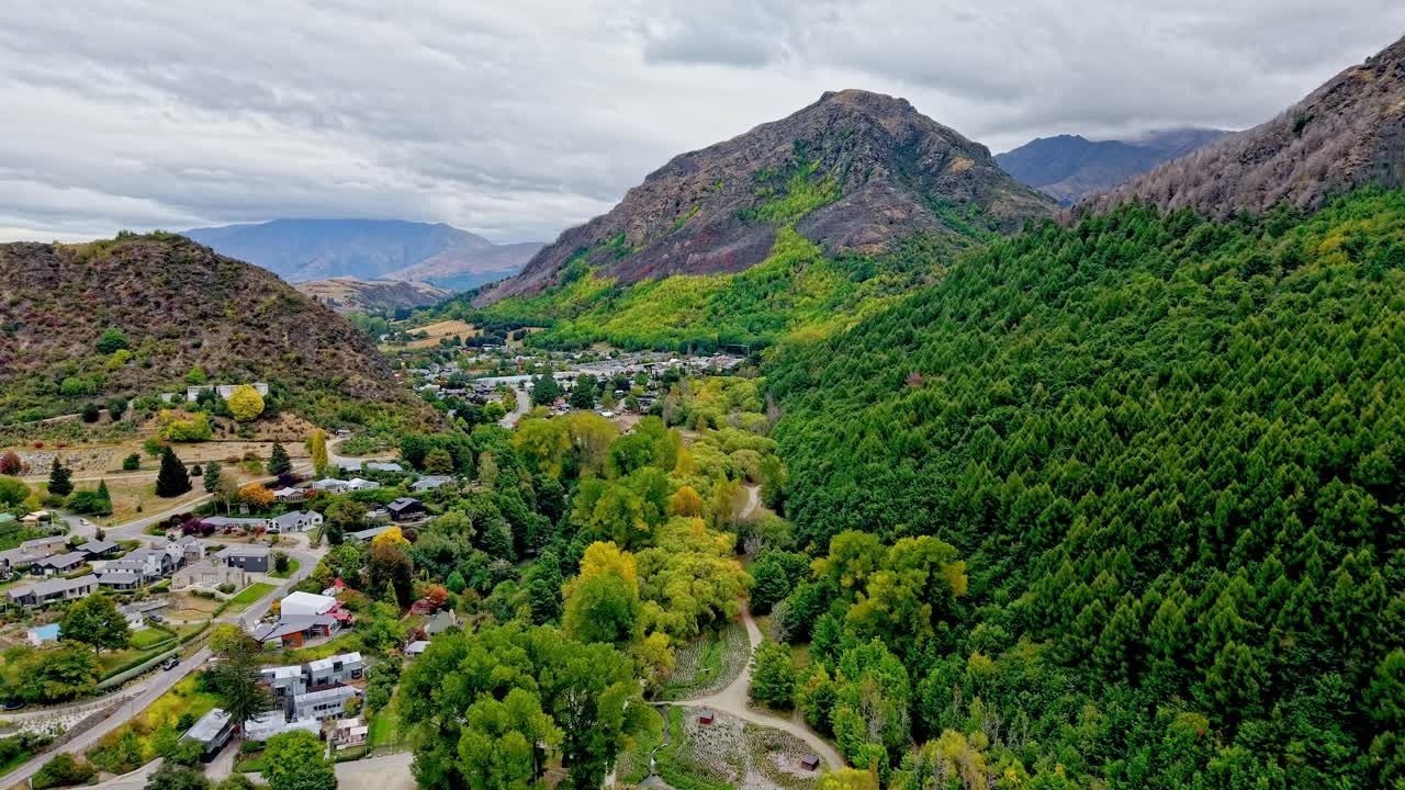 Liten, historisk by i New Zealand viser turstier slukt av levende høstfarger fra et luftperspektiv som flyr mot et nydelig fjellandskap på en skyet dag