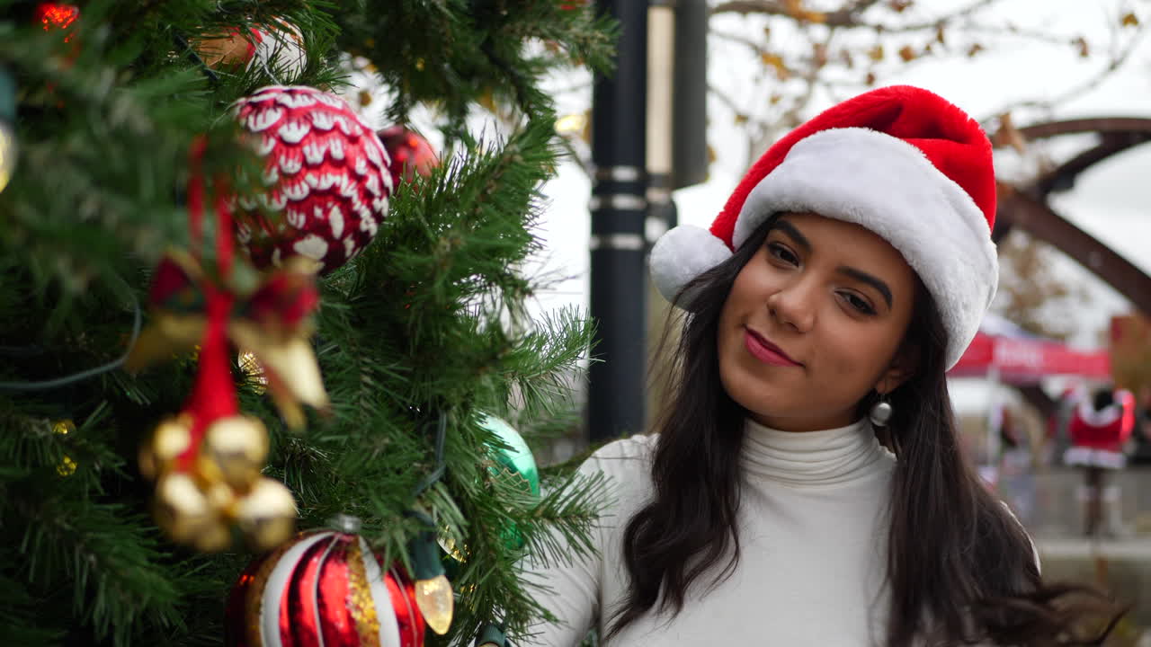 A hispanic woman in a Santa Claus hat looking happy and merry next to a holiday Christmas tree with ornaments and lights