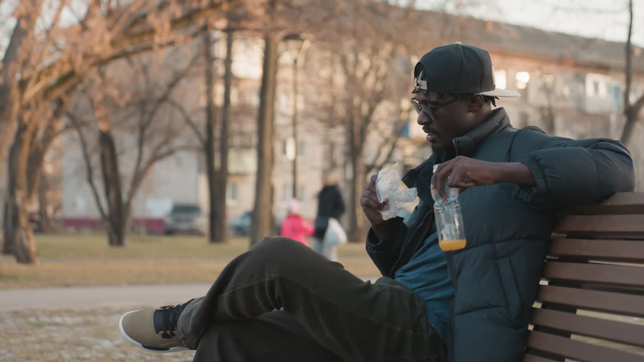 Sidebillede af mørkhåret mand, der sidder på en parkbænk og holder et glas juice og spiser en snack, mens han observerer omgivelserne på en rolig eftermiddag med en sløret baggrund af gående mennesker og bygninger
