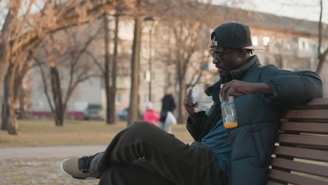side view of dark skinned man sitting on park bench holding jar of juice and eating snack while observing surroundings during calm afternoon with blurred background of people walking and buildings