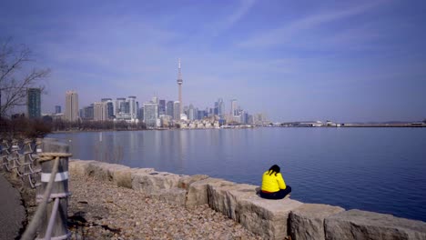 person in toronto trillium park with headphones sitting near lake in bright yellow jacket during day