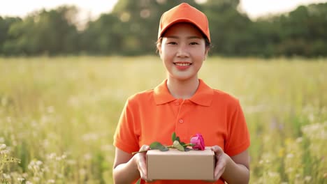 a cheerful young woman wearing an orange outfit stands in a blooming field, smiling while holding a gift box adorned with flowers. the bright sun shines overhead, creating a vibrant atmosphere.
