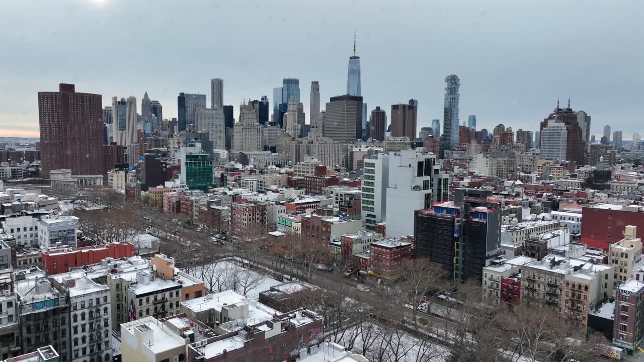 Luftfoto af New York City om vinteren, der viser et tæt byområde med sneklædte tage, gader og træer uden blade. Manhattans skyline rejser sig i baggrunden under en overskyet himmel. Bredt billede.