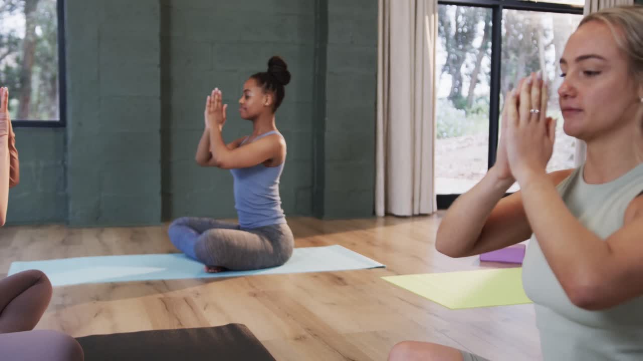Donne di diverse etnie concentrate che meditano insieme sui tappetini in una lezione di yoga con istruttrice, al rallentatore