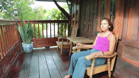 Thailändische-Dame-Sitzt-Draußen-Auf-Dem-Balkon-Eines-Traditionellen-Thailändischen-Hauses-Und-Bewundert-Die-Natur-In-Ayutthaya,-Thailand