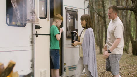 feliz madre vertiendo té de la tetera en la taza de su hijo mientras está de pie junto a la autocaravana en el bosque