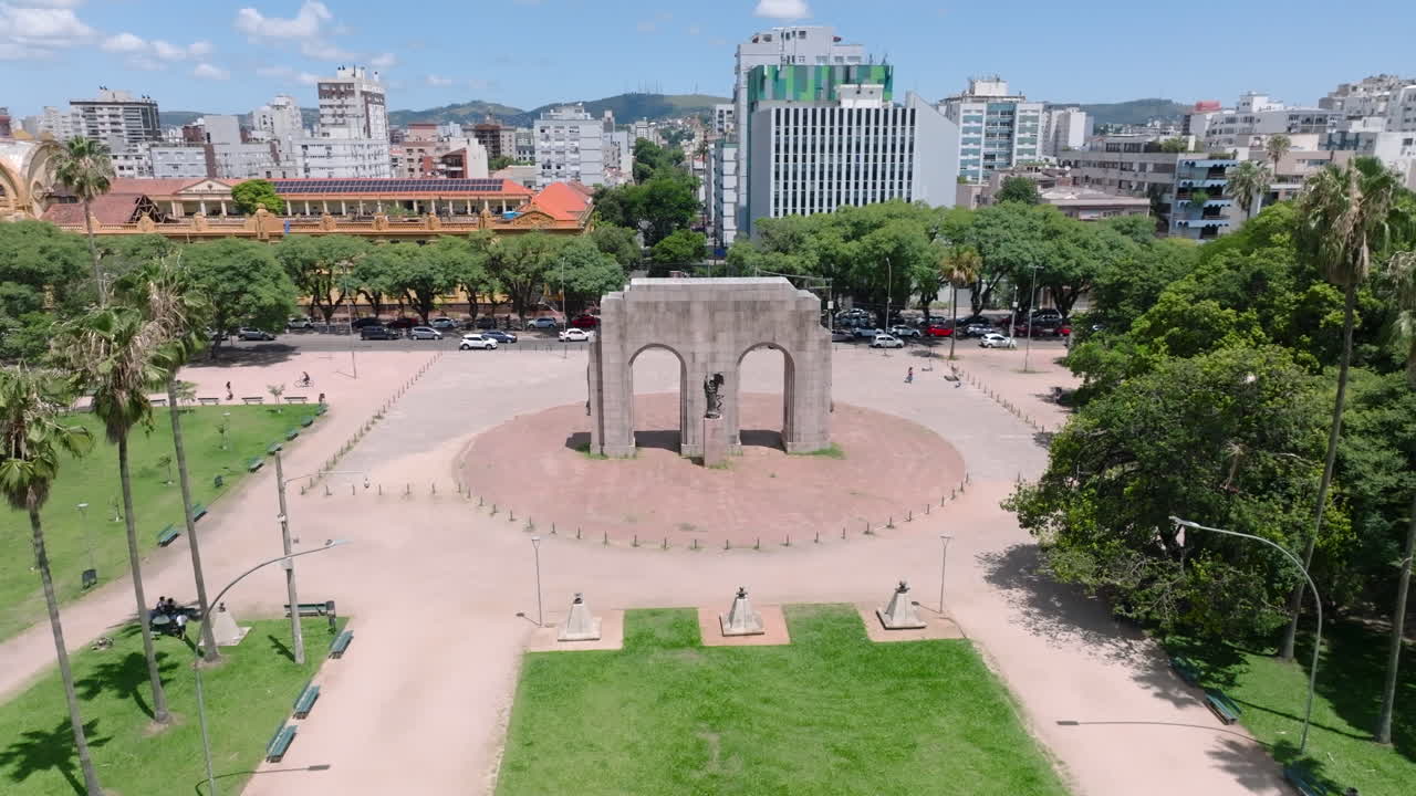 Sorvolo aereo cinematografico con drone che rivela l'arco del Monumento ao Expedicionario dietro le palme nel Parco Farroupilha. Punto di riferimento storico a Porto Alegre. Video 4K D-Log a 10 bit