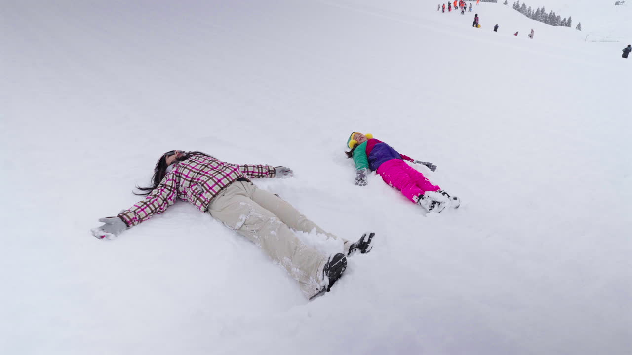 Static view shows an adult and child lying side by side in fresh snow, creating snow angels with their outstretched limbs and colorful winter clothing, capturing a playful and intimate winter moment