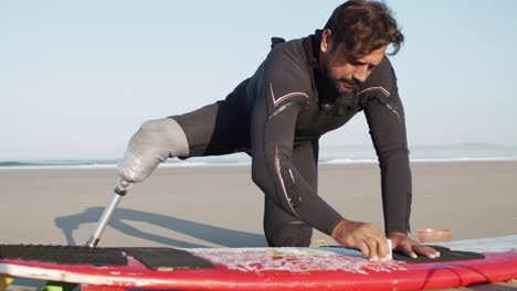 static shot of a male surfer with prosthetic leg waxing surface of surfboard