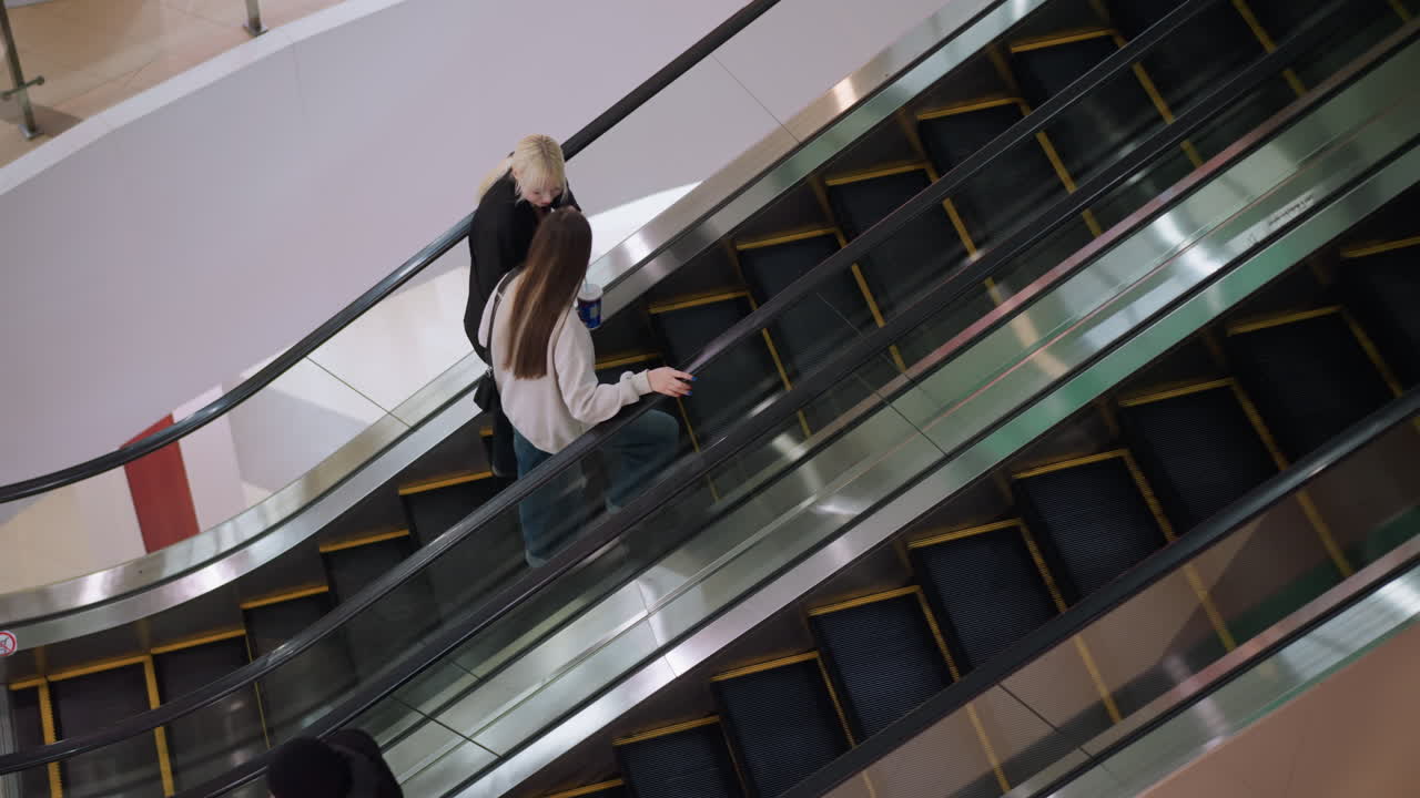 Ovenfra og ned-perspektiv af to venner, der træder op på en rullende trappe, mens de holder en kop med drikkevare og snakker, mens en anden person går ned på den modsatte side, hvilket viser en afslappet interaktion