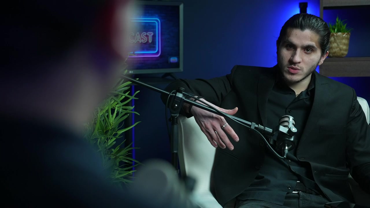 Ung mandlig podcaster i en sort blazer kigger og taler direkte til værten under en interviewoptagelsessession i et moderne studie, indholdsskabelse og mediekoncepter