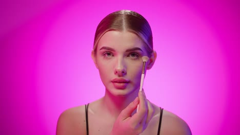 young caucasian blonde woman brushes her eyelids with dusting powder makeup studio closeup portrait shot, pink background