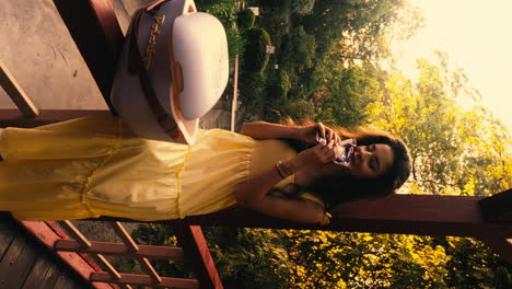 slow motion dolly shot of a young woman in a yellow dress taking a bite of a delicious bar of chocolate and enjoying it on the wooden terrace while her picnic bag stands in front of her