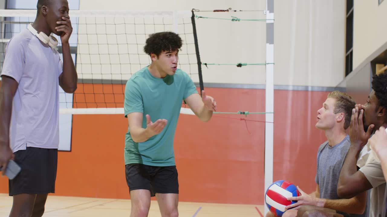 体育館の中央で、多様な男性チームの選手がチームメイトに話しかけ、ボールで手本を示し、スマートフォンを確認している。