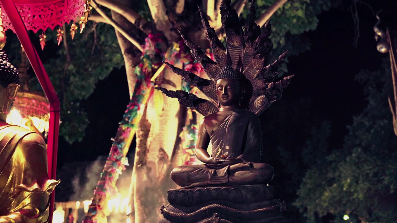 Static medium shot of a Phra Nak Prok bronze Buddha in meditation pose sheltered by nine-headed Naga serpent with colorful Yi Peng garlands sacred Bodhi tree, and drifting mist at Northern Thai temple
