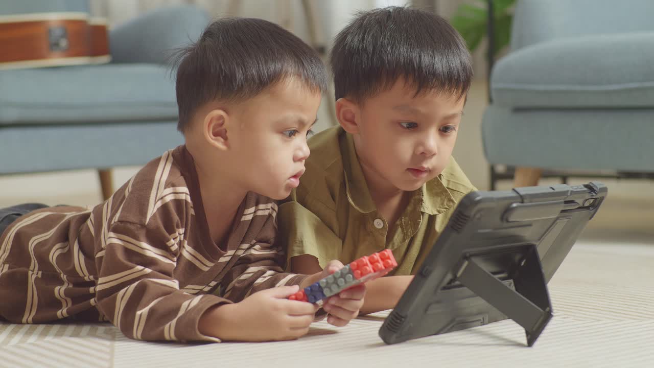 플라스틱 장난감 벽돌을 가진 아시아 어린이들이 바닥에 누워서 집에서 함께 태블릿에서 영화를 보고 있습니다.