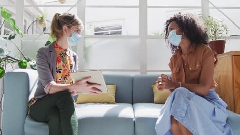 Two-woman-wearing-face-mask-discussing-over-digital-tablet-at-office