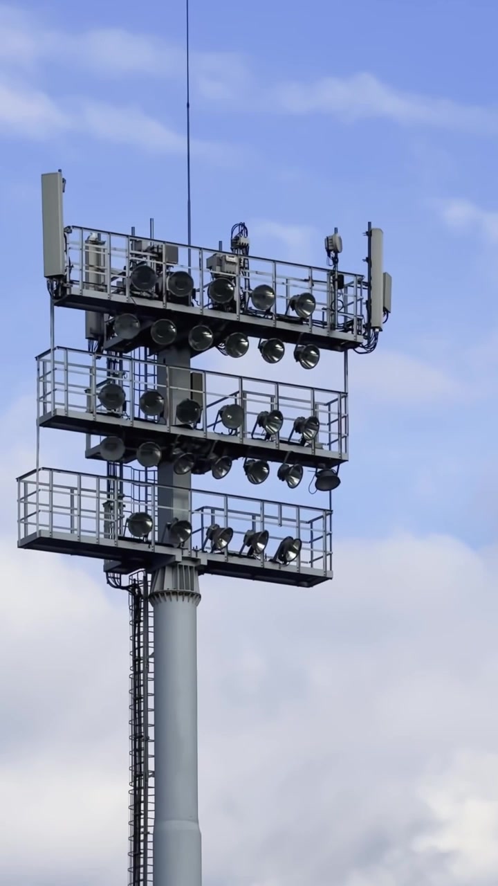 Primo piano verticale di una torre faro da stadio con antenne per telecomunicazioni, piattaforme di sicurezza e lampade montate contro un cielo blu nuvoloso.