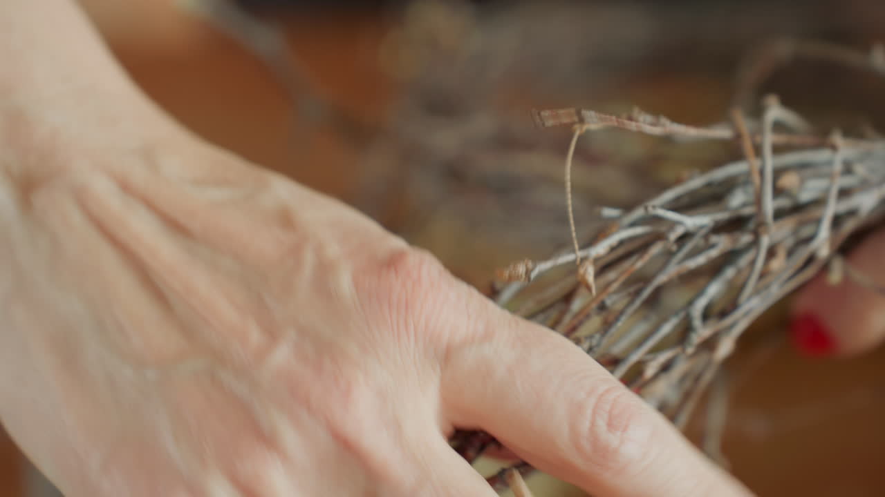 Female florist hands with red nails assembling natural twig wreath on wooden table, showing close up of detailed crafting process, rustic materials, and seasonal handmade decorative arrangement
