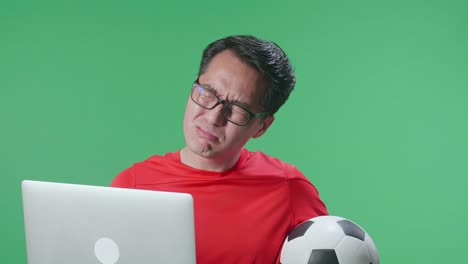 vista lateral de cerca de un hombre asiático con una pelota se arrepiente de animar el fútbol en una computadora portátil en el estudio de pantalla verde