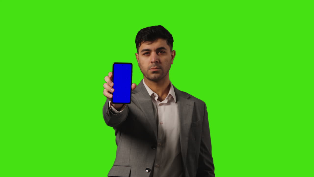 진지 한 사업가 가 녹색 스크린 배경 에 서 있는 카메라 를 향해 파란 스크린 휴대 전화 를 들고 있다
