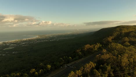 layers of mist rise through illawarra escarpment forested slopes at sunrise in australia with cars driving along road, aerial panoramic view to town below