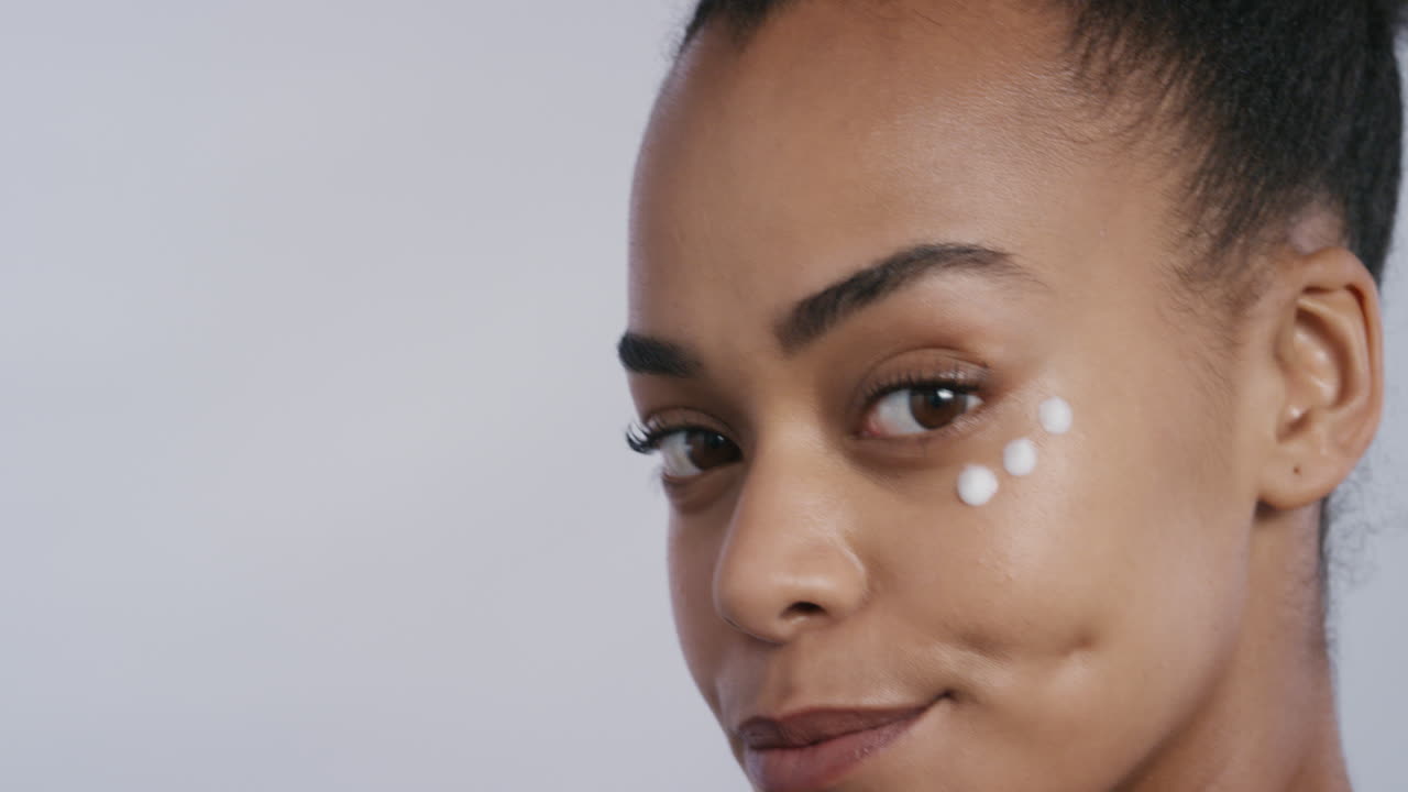 ritratto bella giovane donna afroamericana che applica crema idratante per la pelle sul viso godendo dell'esfoliazione massaggiando lozione anti invecchiamento di lusso bellezza femminile perfetta su sfondo bianco concetto di cura della pelle